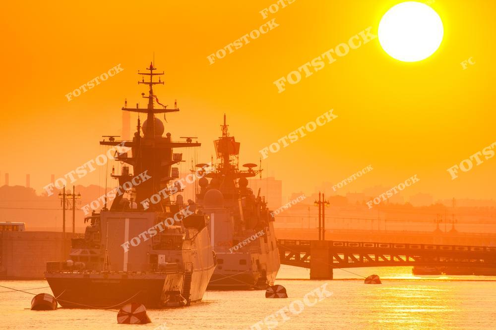 На фотографии изображены военные корабли, стоящие на якоре в водах Санкт-Петербурга, под светом заходящего солнца. Яркий оранжевый закат окрашивает сцену в тёплые тона, подчеркивая силуэты судов и отражение солнечного света на воде. Закатное солнце, почти касающееся горизонта, добавляет драматичности, придавая этому морскому пейзажу особую атмосферу.